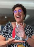 Dr. Fried-Oken holds up a clear glass award with red and white lettering 'isaac the fellowship award' wearing a black and white top, and a wide open smile that reaches her eyes behind red rimmed glasses that match the red accent. 